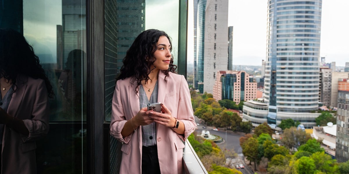 professional woman standing outside on balcony, holding cell phone, looking out 