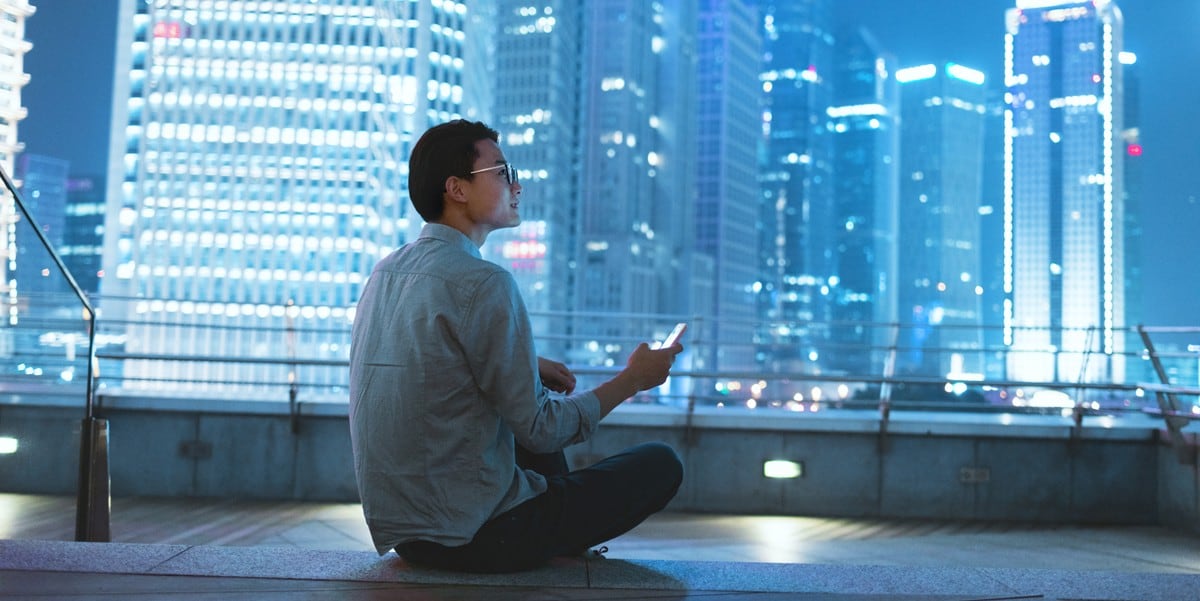 man sitting on rooftop holding cellphone, lit up skyscrapers in front of him