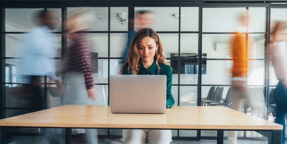 woman working at desk with laptop, in the office, people walking behind her